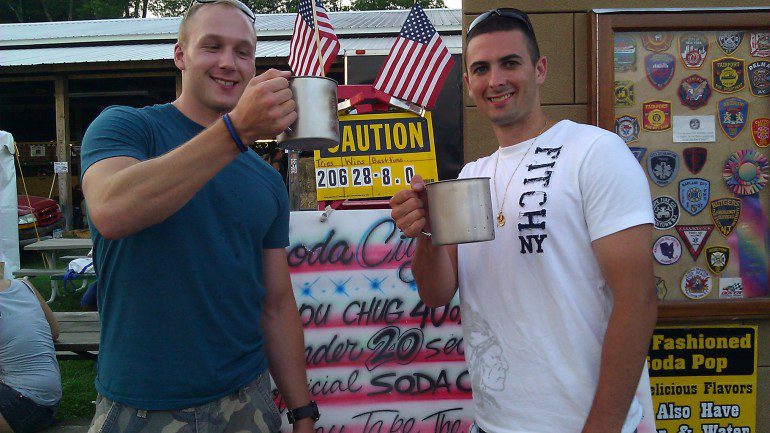 Two men with mugs participating in drinking challenges in front of a sign.