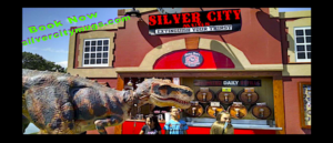 A group of people are standing in front of a t-rex in front of a silver city restaurant.
